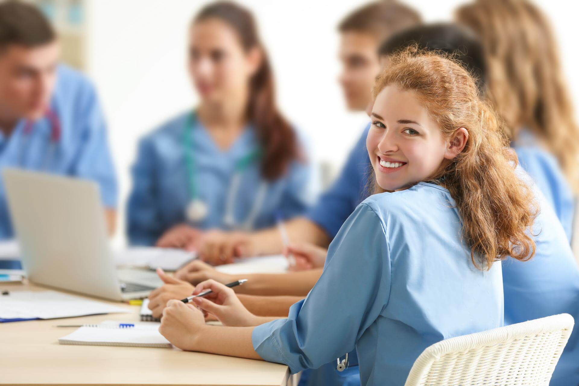 Eine Gruppe von Personen in blauer medizinischer Arbeitskleidung sitzt an einem Tisch und nimmt an einer Besprechung oder Schulung teil. Auf dem Tisch liegen Notizbücher und ein Laptop, während die Gruppe konzentriert arbeitet. Eine Person lächelt freundlich in die Kamera.